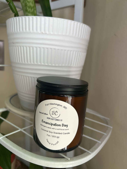 Emancipation Day Coconut soy wax candle in amber jar with black lid on white shelf next to white planter