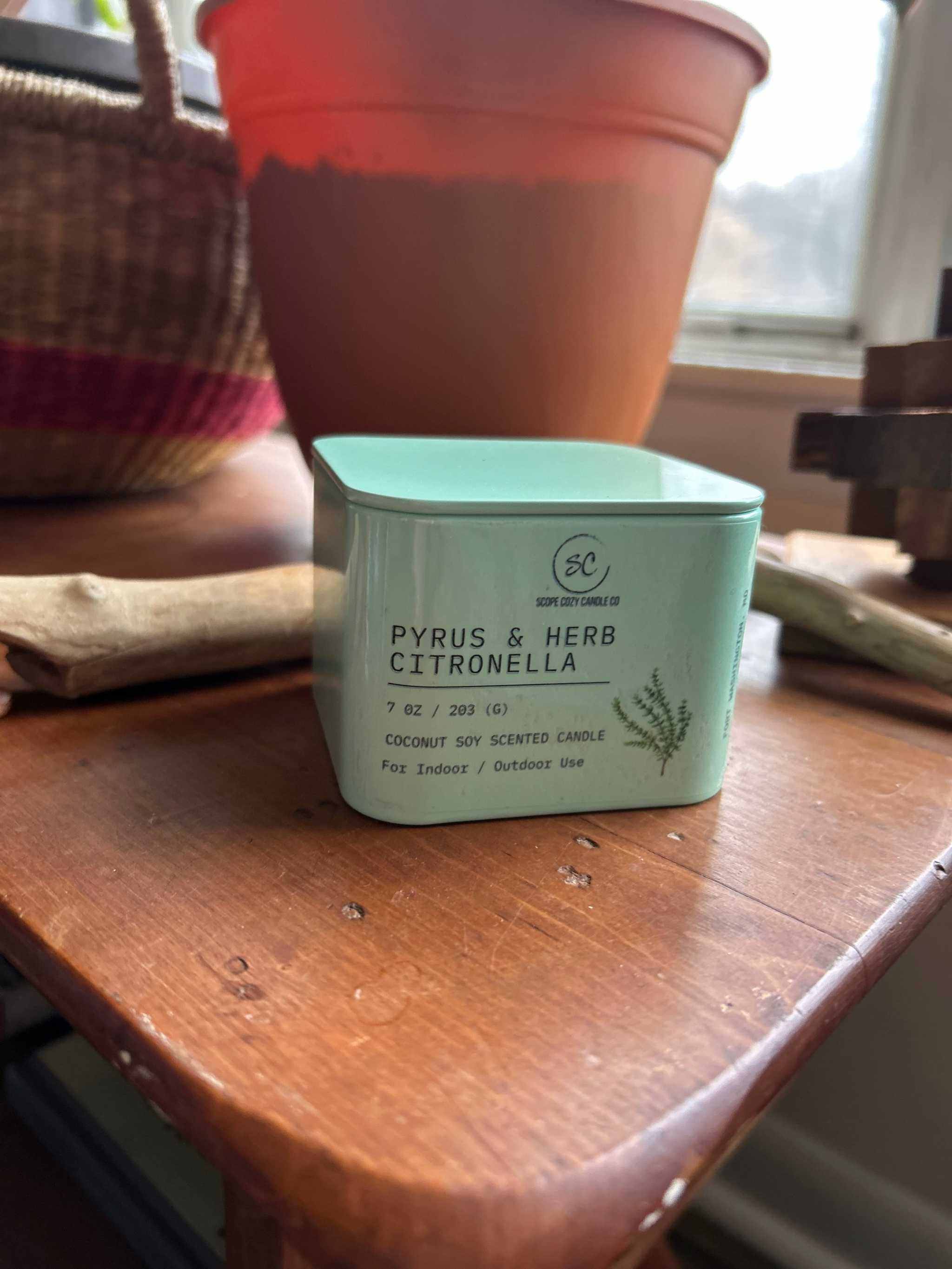 Pyrus &amp; Herb Citronella scented candle in a green square container on a wooden table near a window and plant pot