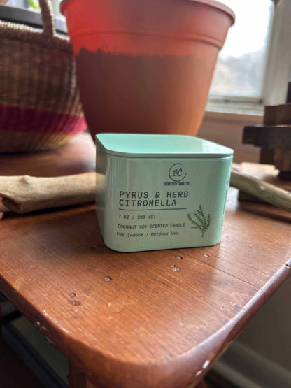 Pyrus &amp; Herb Citronella scented candle in a green square container on a wooden table near a window and plant pot