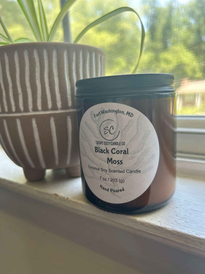 Black Coral Moss coconut soy candle in amber glass jar with black lid on windowsill next to plant pot