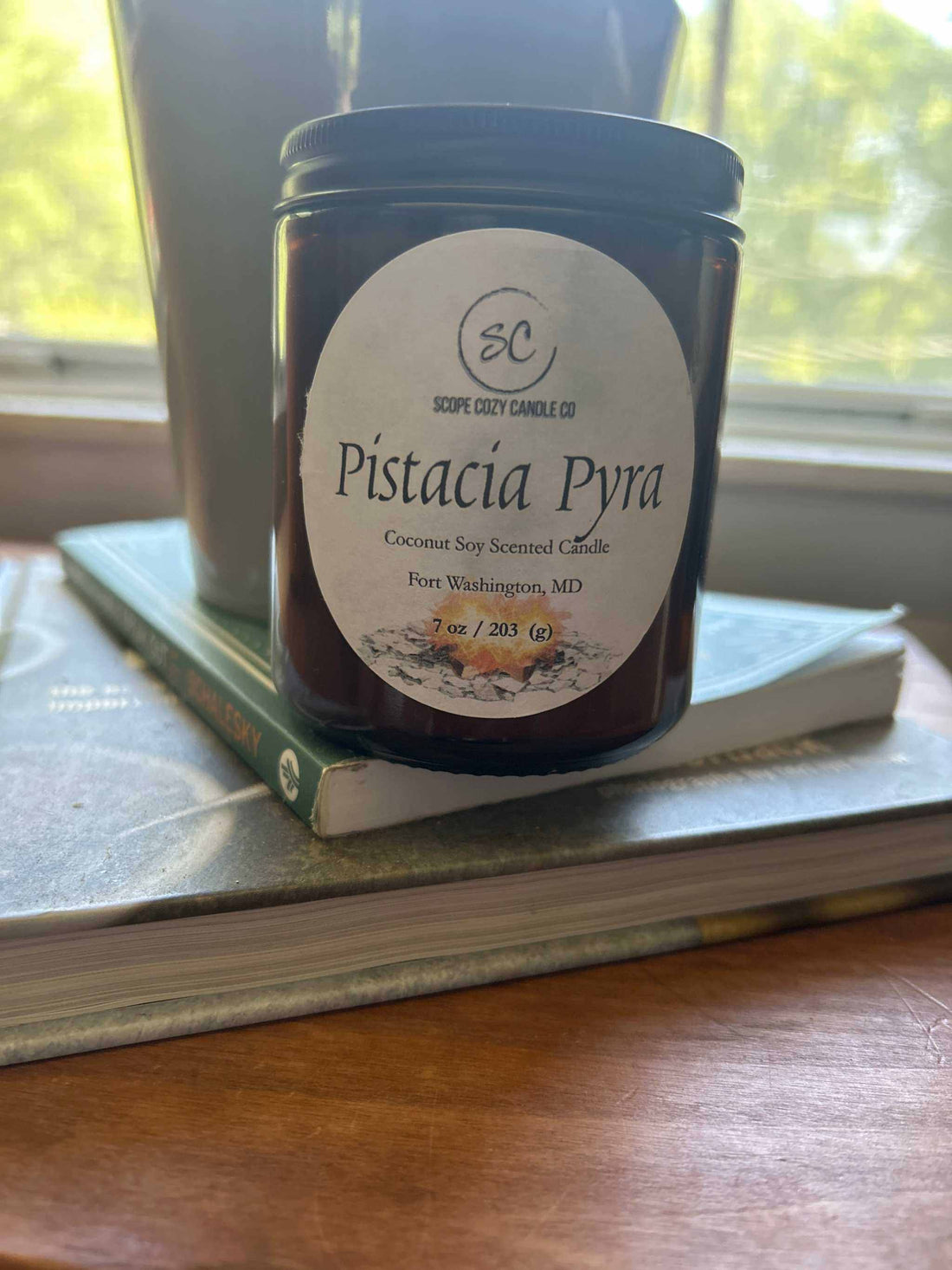 Pistacia Pyra Coconut Soy scented candle in amber glass jar on books by window