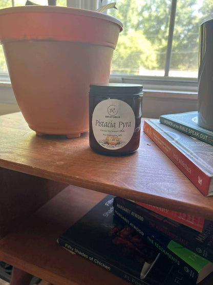 Pistacia Pyra Coconut Soy scented candle in black jar on wooden shelf near plant and books