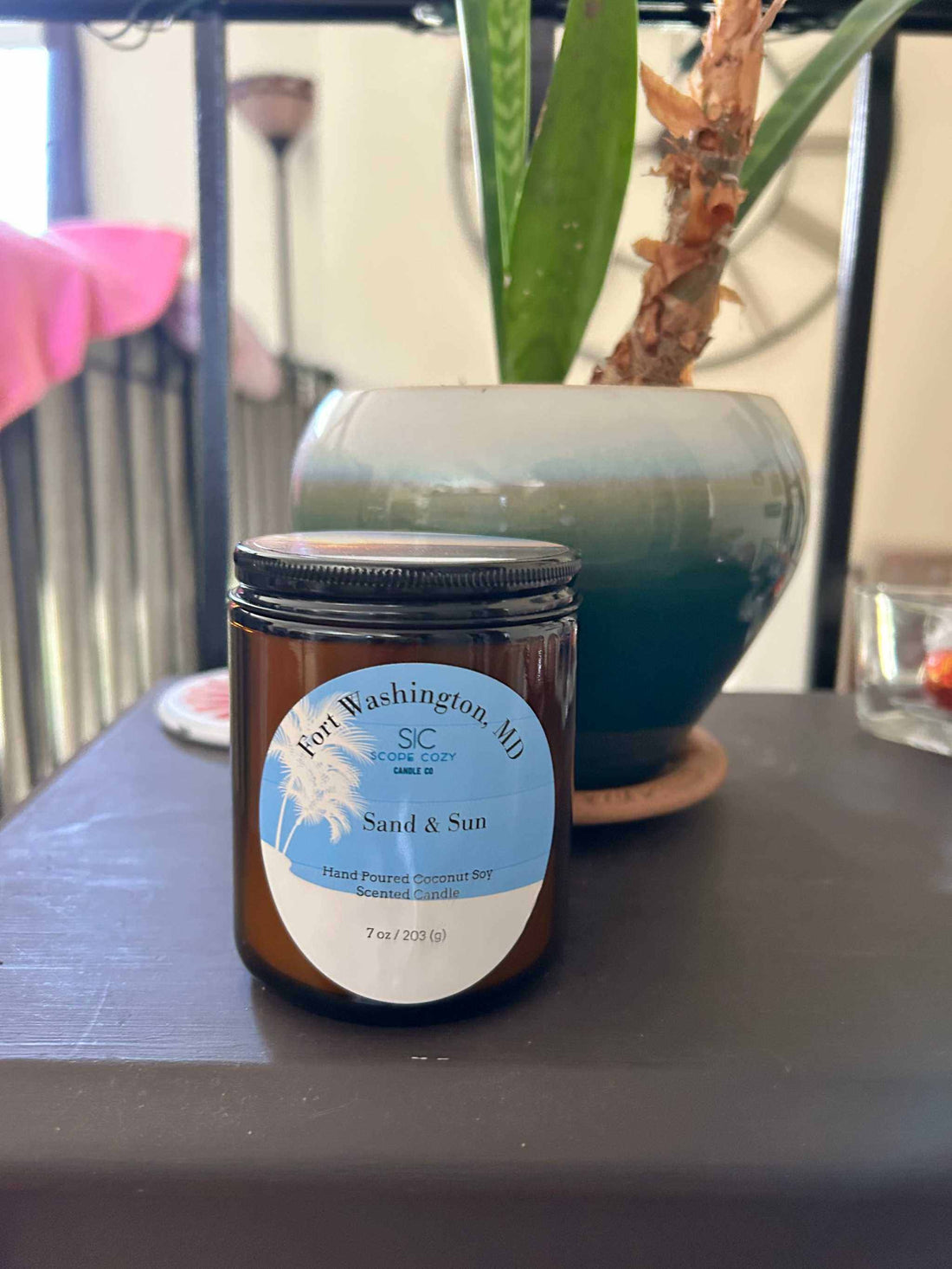 7 oz Sand &amp; Sun Coconut soy scented candle in amber jar with black lid on table near green potted plant